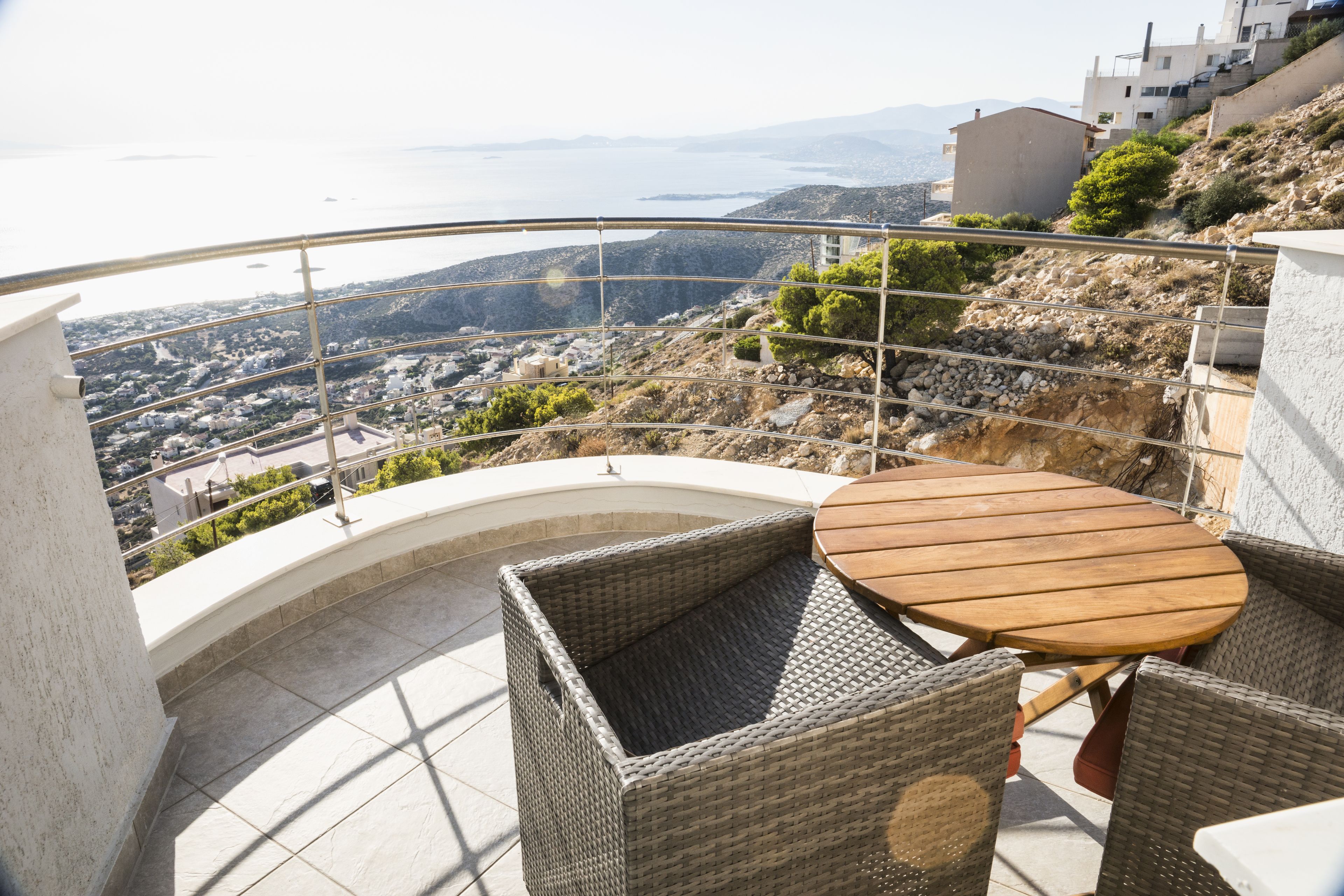 Un balcón con mobiliario de mimbre, una mesa redonda y vistas a un pueblo costero y al mar.