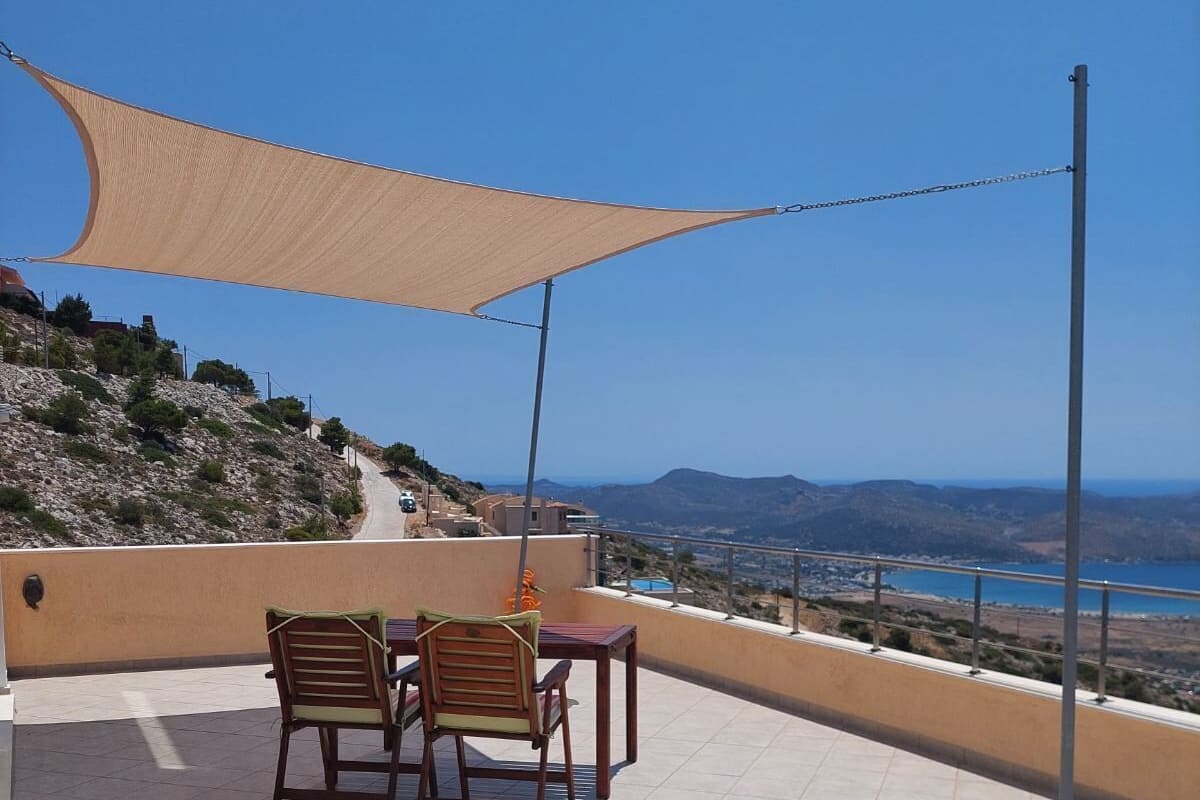 A terrace with two wooden chairs and a table, a beige shade sail, and a view of a coastal landscape.