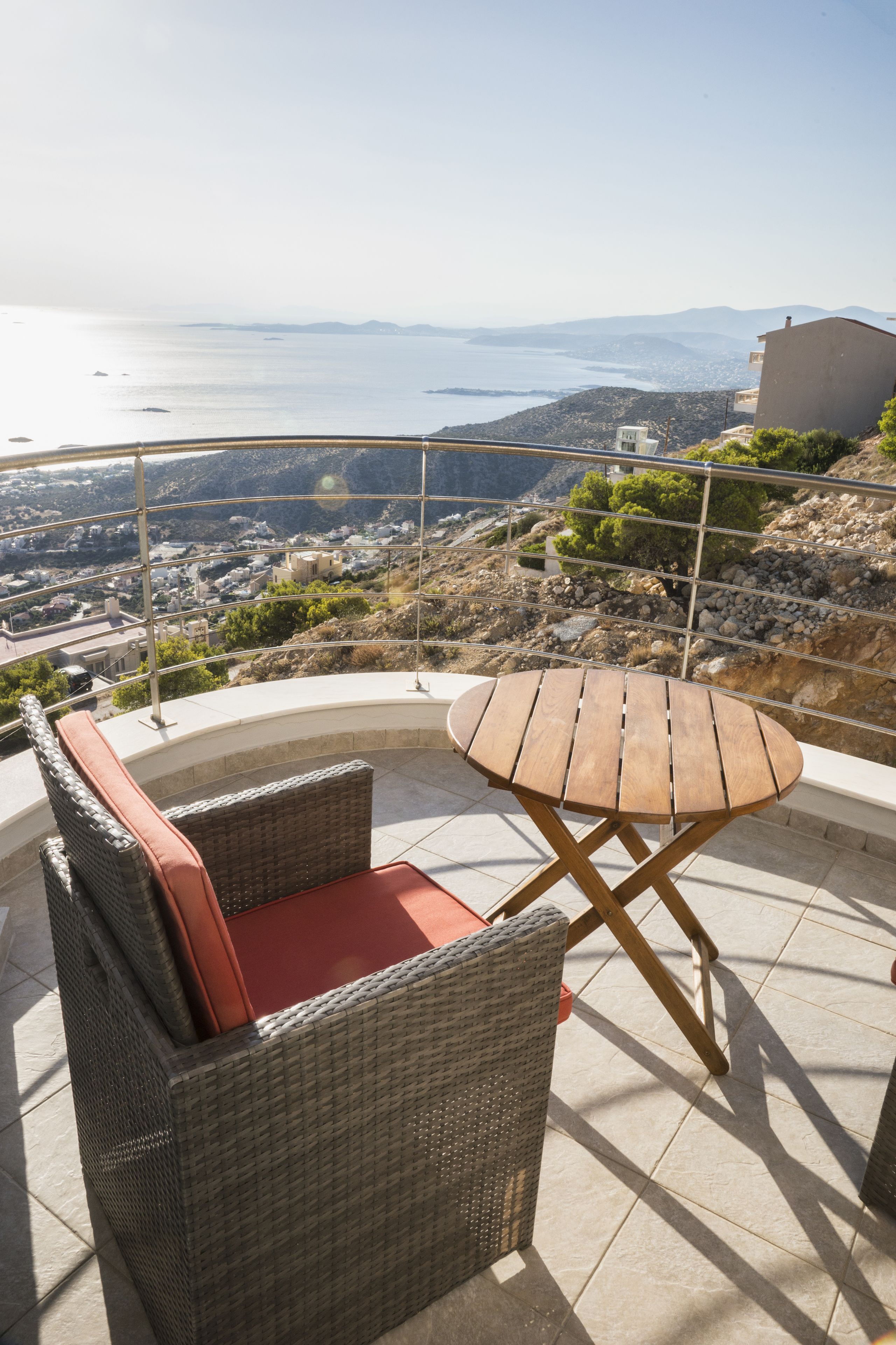 Une chaise en osier avec un coussin rouge et une table en bois sur un balcon donnant sur un paysage urbain côtier.