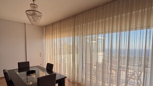A spacious room with a dining table, chairs, a chandelier, and large windows with sheer curtains. - Viva B&B (Saronikos)