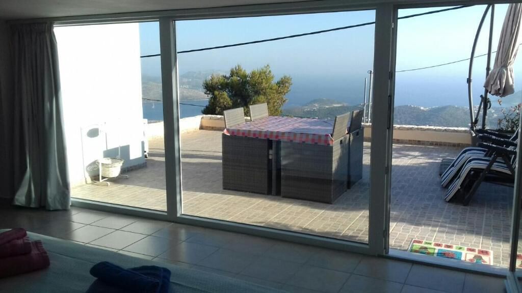 Una habitación con un gran ventanal corredera que da a un balcón con mesa y sillas, vista al mar y una planta en maceta.