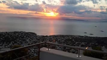 Vista do pôr do sol a partir de uma varanda com vista para a costa rochosa, mar calmo e barcos ao longe.