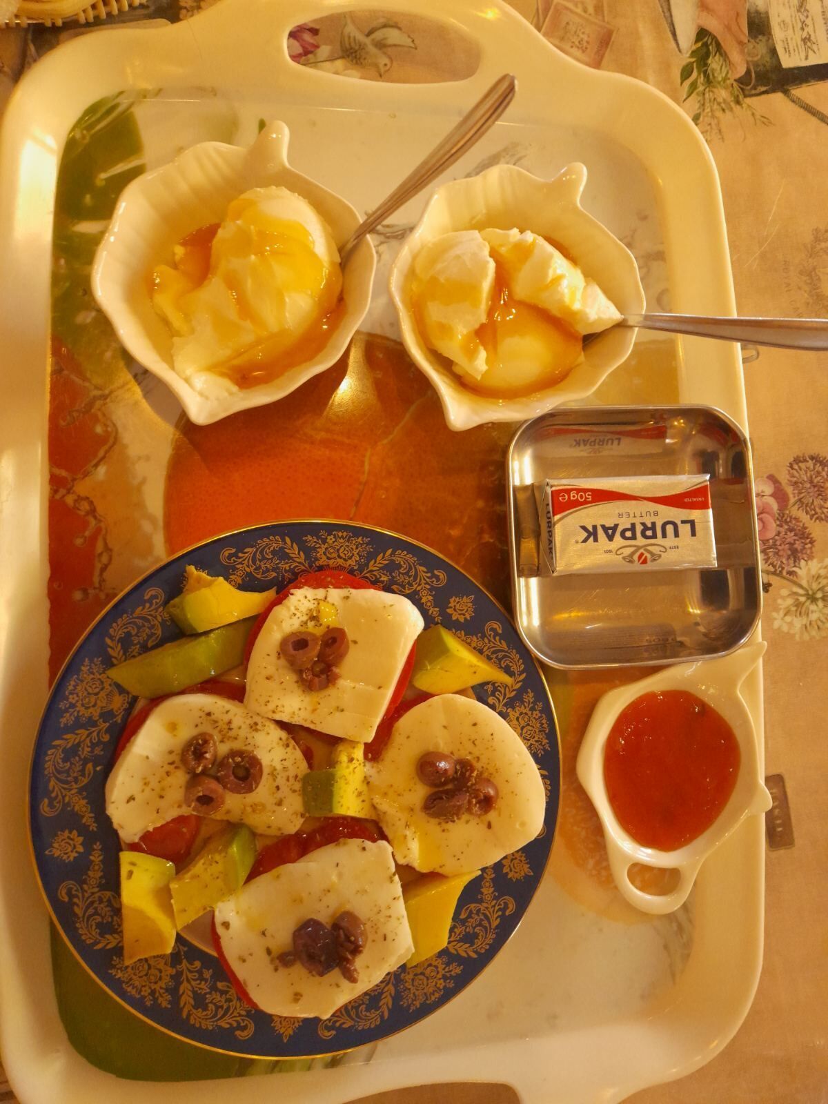 A tray with food items including a plate of sliced vegetables and cheese, two scallop shells with a sauce, a small container of butter, and a cup of red sauce.