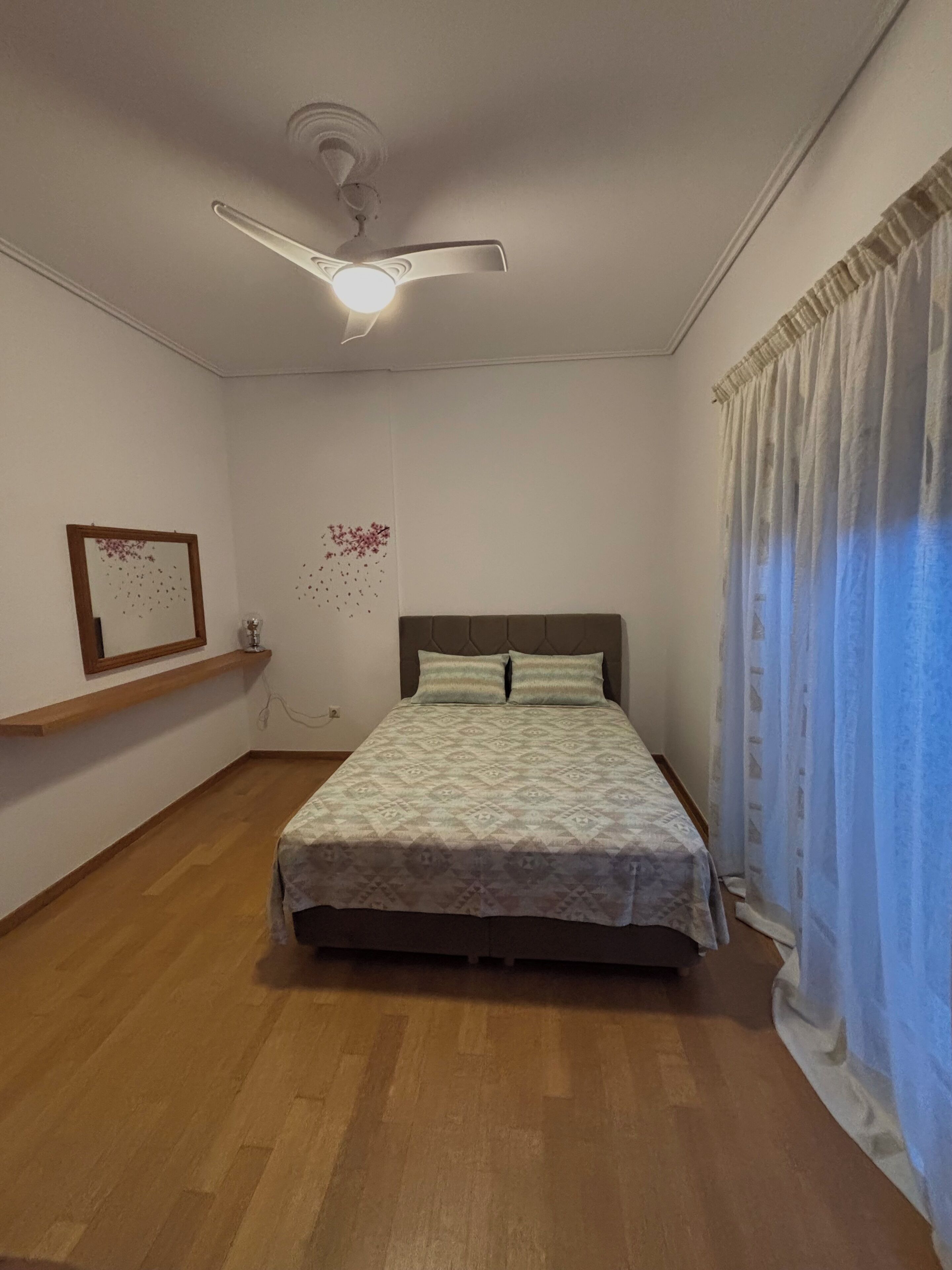A bedroom with a bed, a ceiling fan, a mirror, a shelf, and a window with curtains.