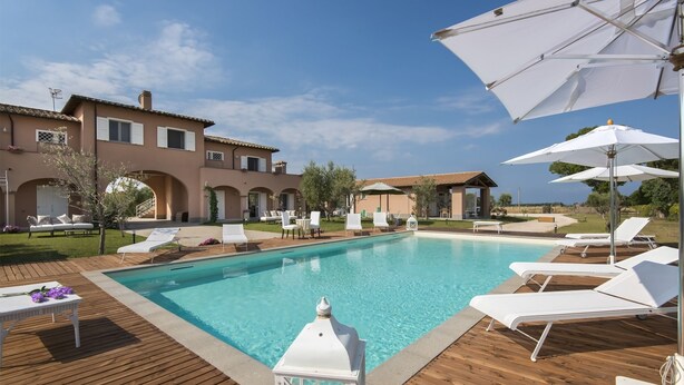 A swimming pool with lounge chairs and umbrellas, surrounded by a wooden deck and a building with arches. - Villa Marina Velca (Tarquinia)