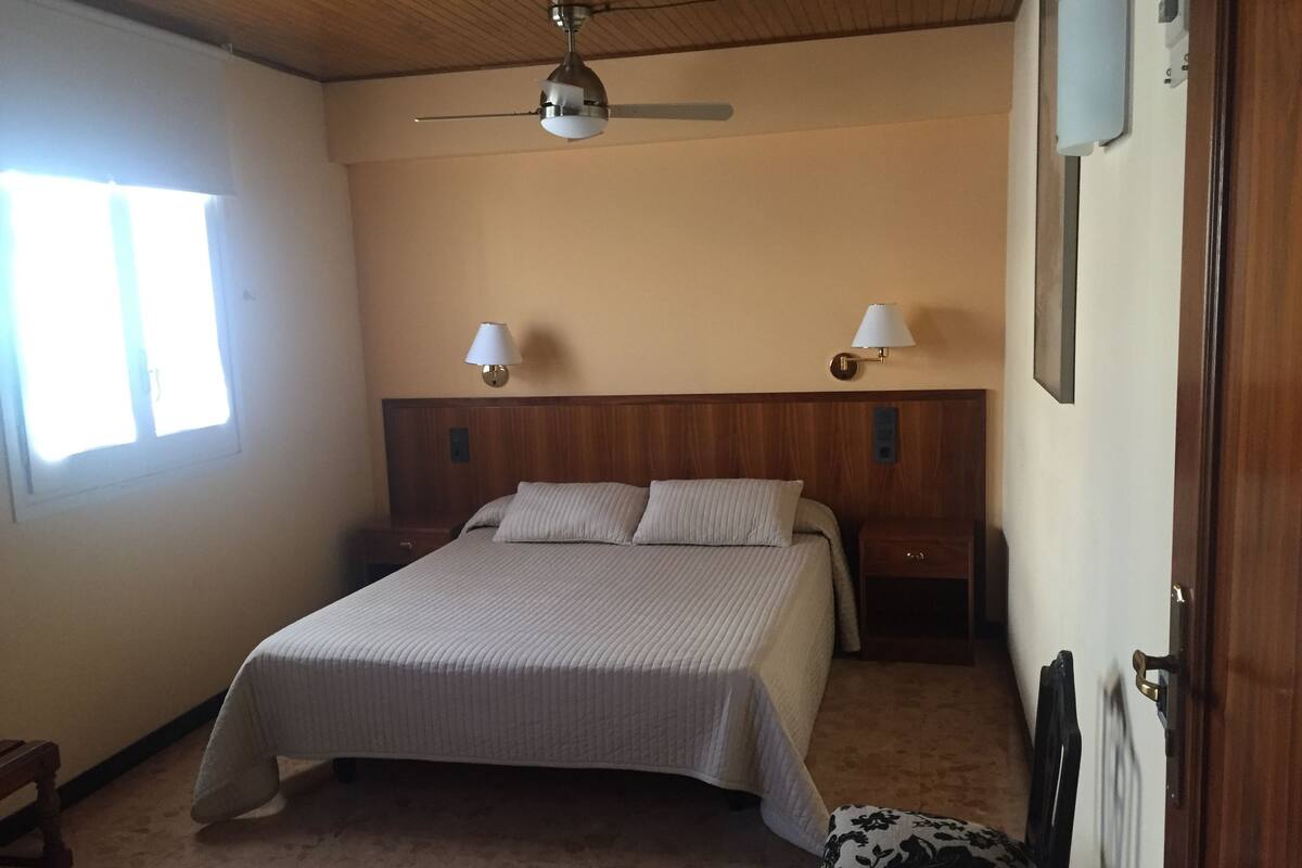 A bedroom with a large bed, two bedside tables, a ceiling fan, and two wall-mounted lamps.