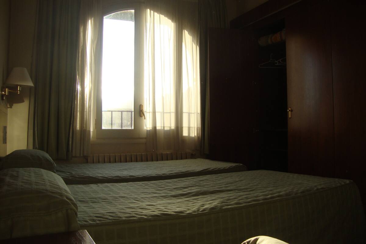 A room with two beds, a window with curtains, a lamp, and a wardrobe.