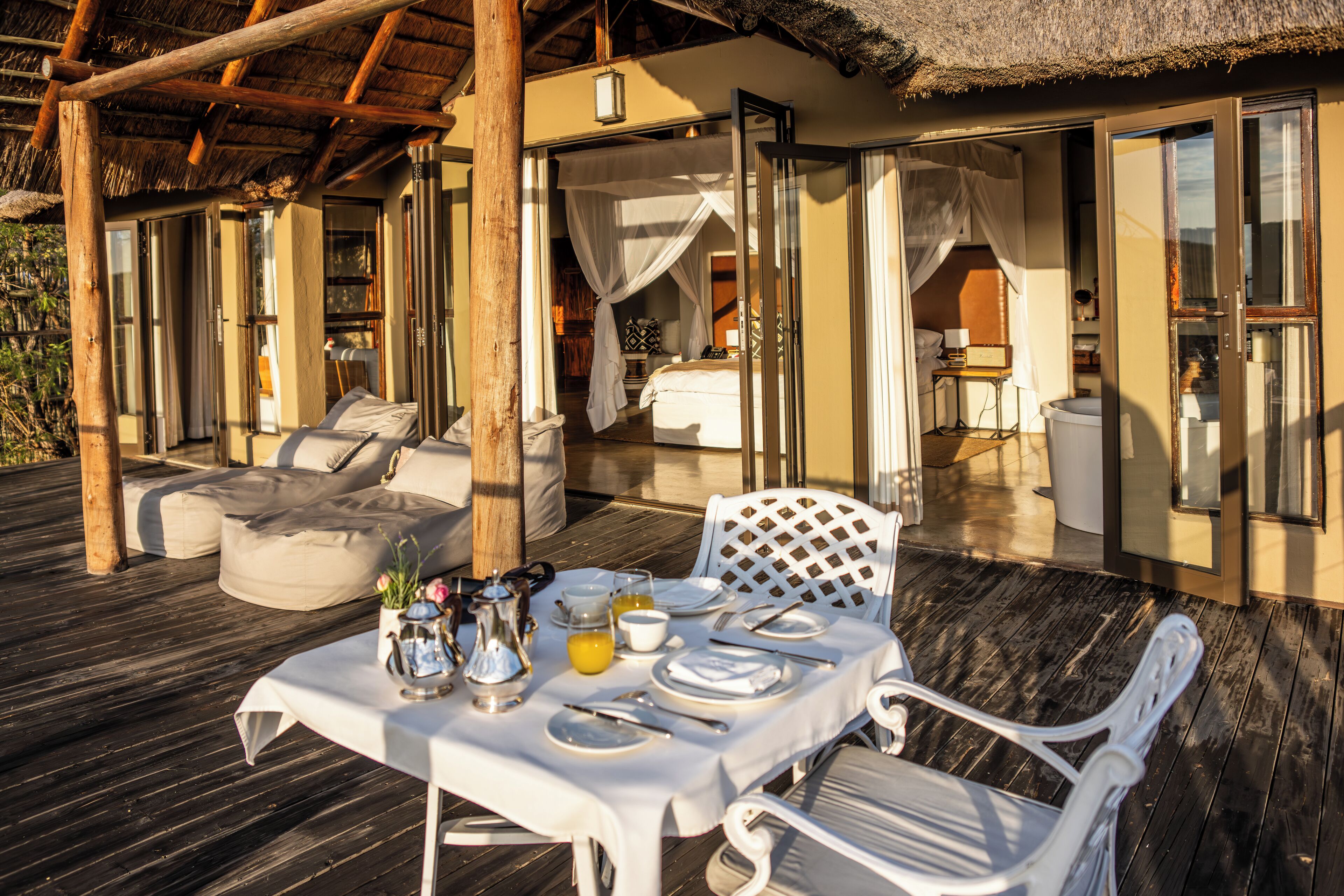 An outdoor dining area with a table set for two, a cushioned seating area, and a thatched roof structure.