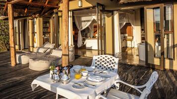 An outdoor dining area with a table set for two, a cushioned seating area, and a thatched roof structure.