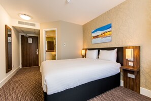 A hotel room with a large bed, a wooden headboard, a painting on the wall, and a carpeted floor.
