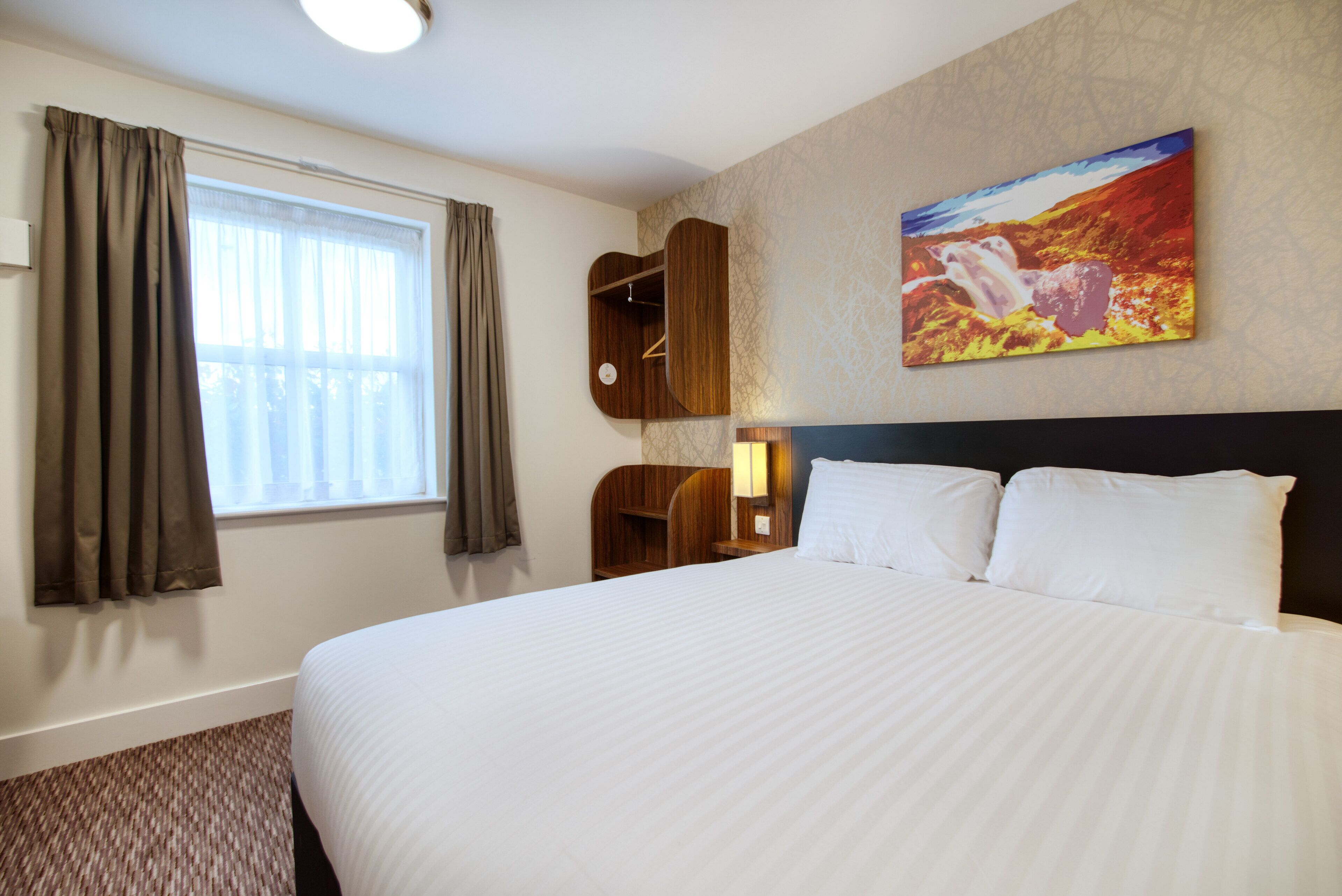 A hotel room with a large bed, a window with curtains, a wall-mounted shelf, and a painting.