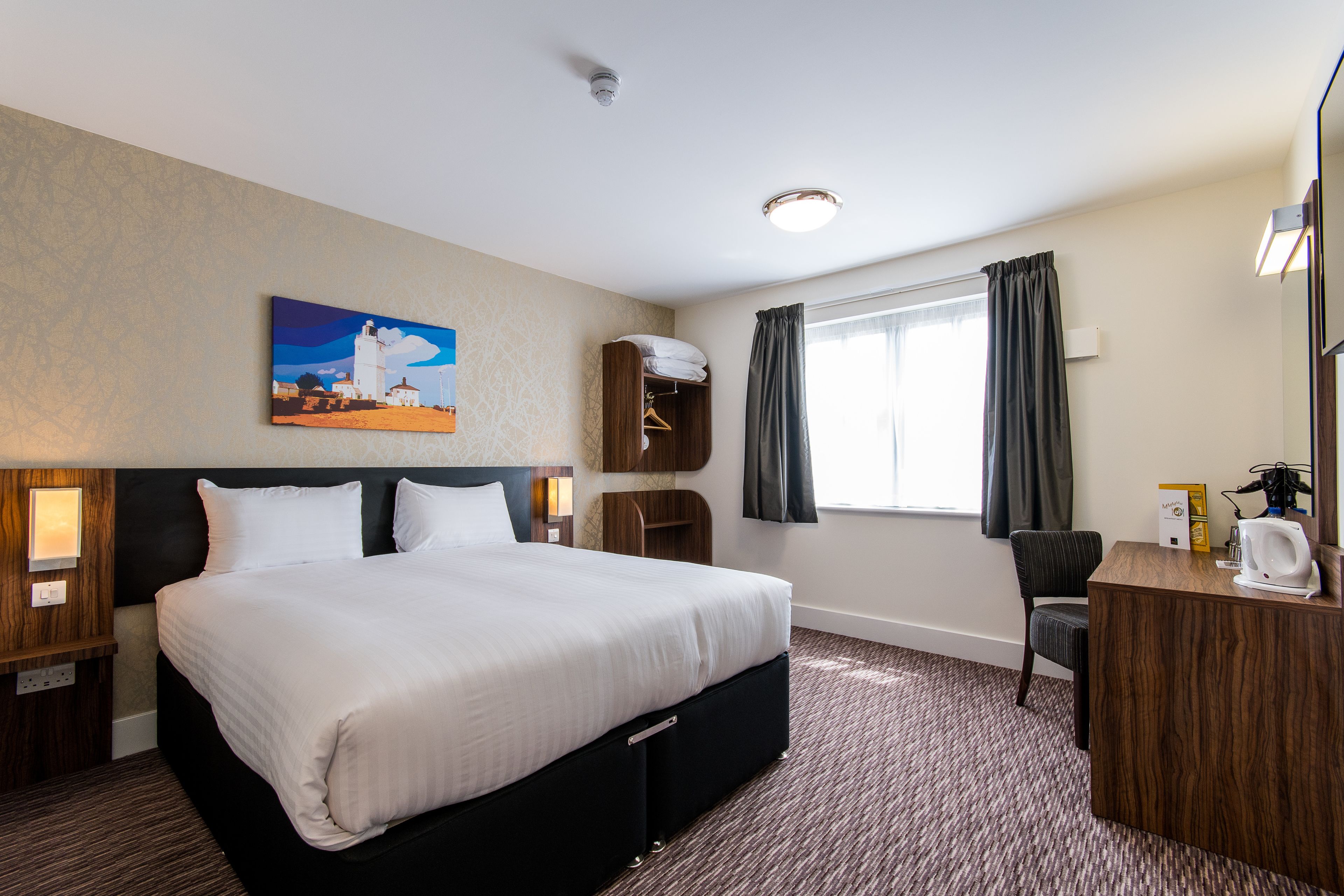 A hotel room with a large bed, a desk with a coffee maker, a chair, a window with curtains, and a painting on the wall.