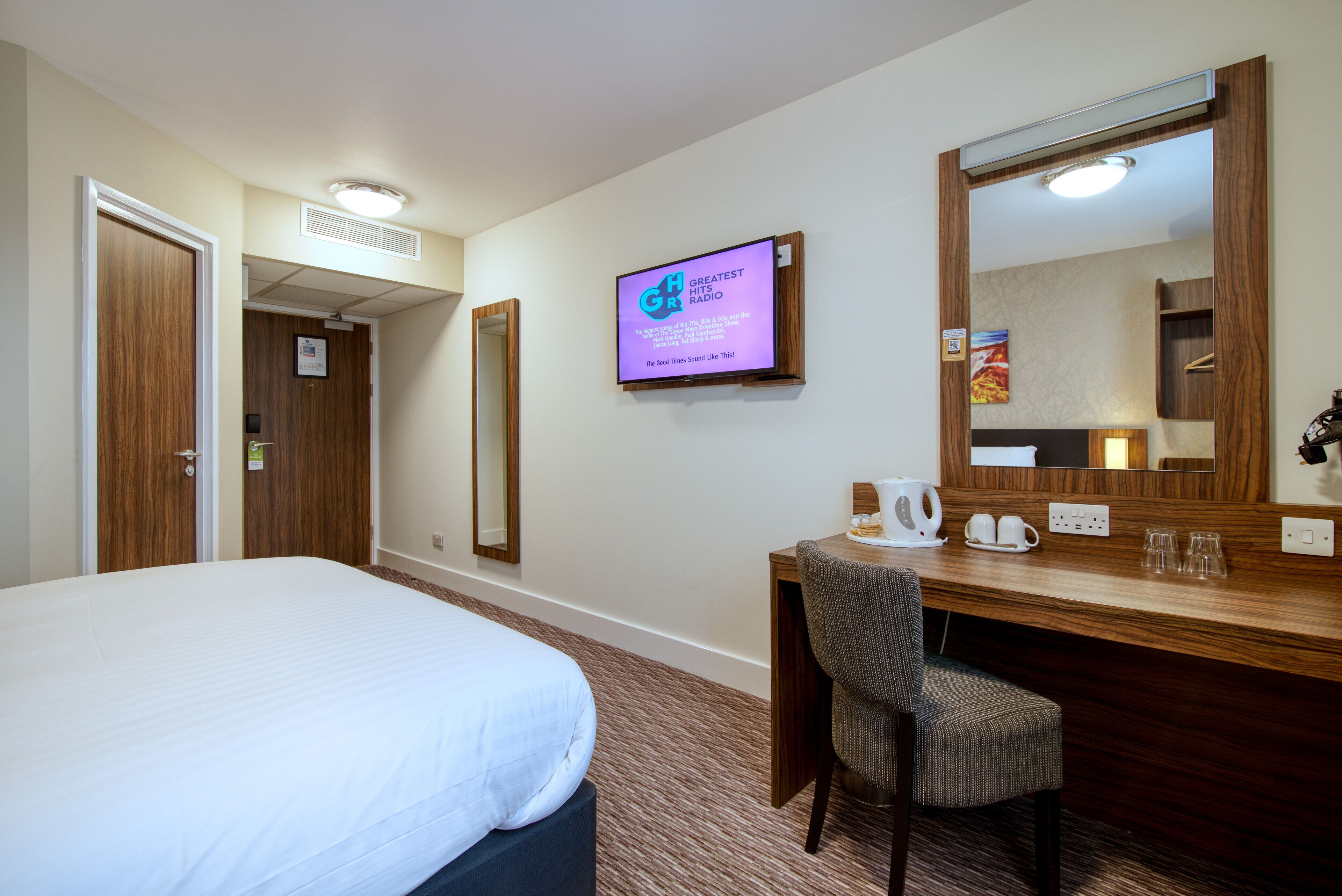 Habitación de hotel con cama, escritorio con silla, televisor mostrando 'Greatest Hits Radio' y un espejo grande.