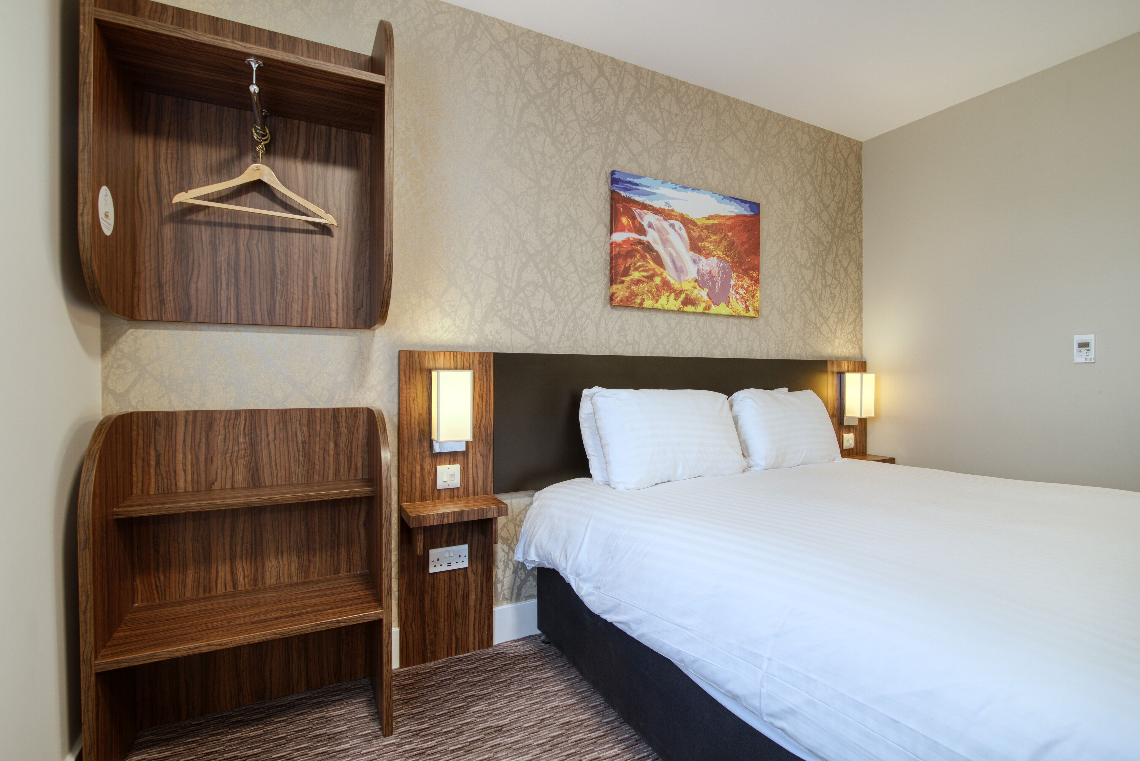 A hotel room with a large bed, wooden headboard, a hanging coat rack, a painting of a waterfall, and bedside lamps.