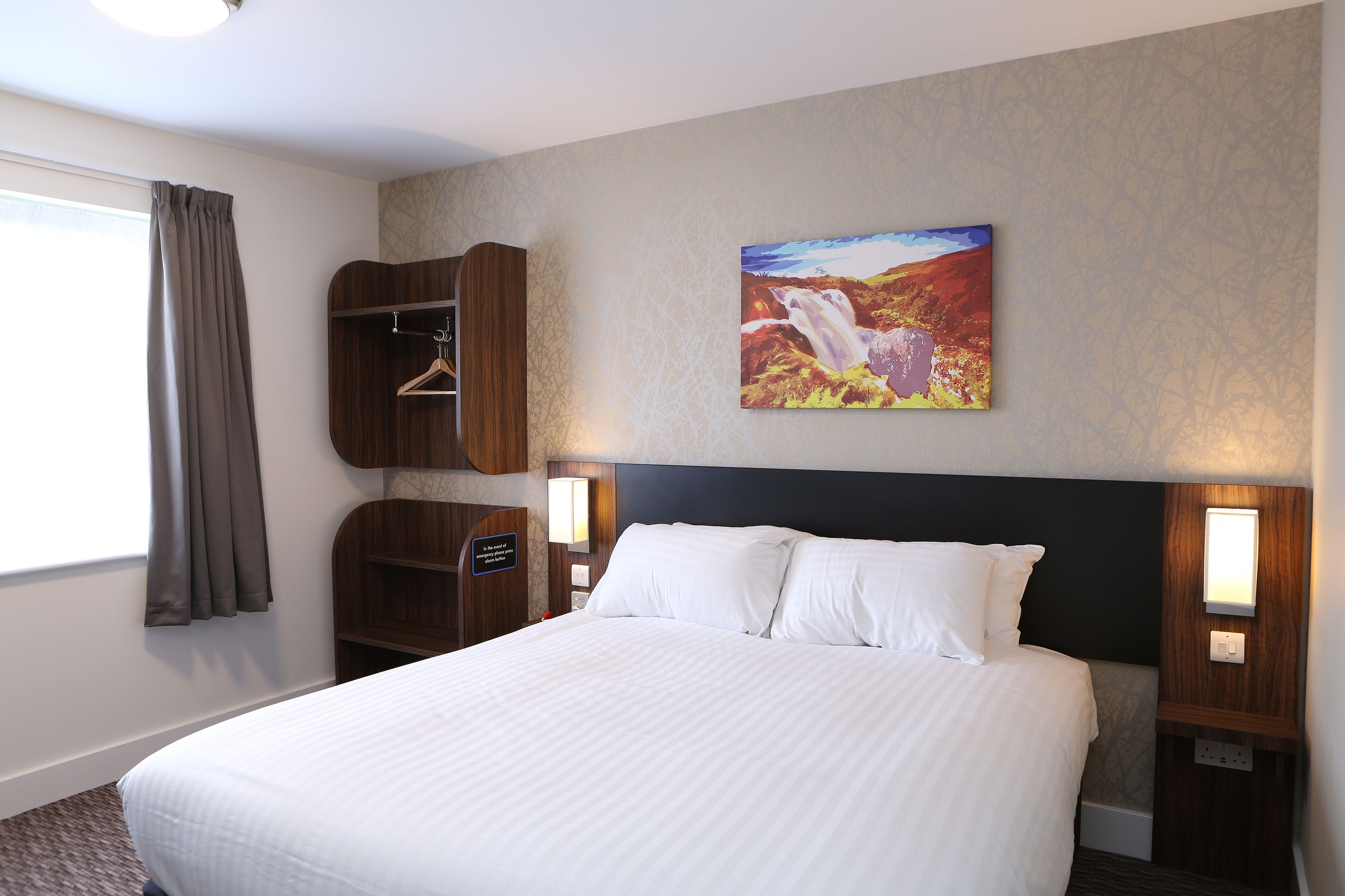Habitación de hotel con una cama grande, un armario, un cuadro y una mesita de noche con una lámpara.