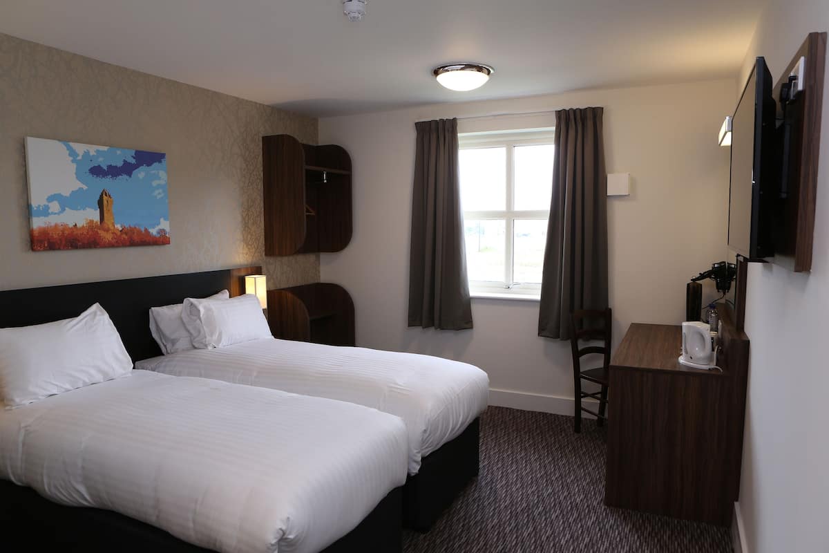A hotel room with two beds, a desk, a chair, a painting, and a window with curtains.