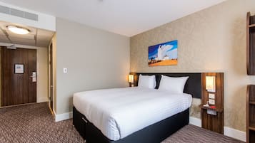 Une chambre d’hôtel avec un grand lit, une tête de lit dotée d’un luminaire, un tableau représentant un phare et une table de chevet en bois avec une lampe.