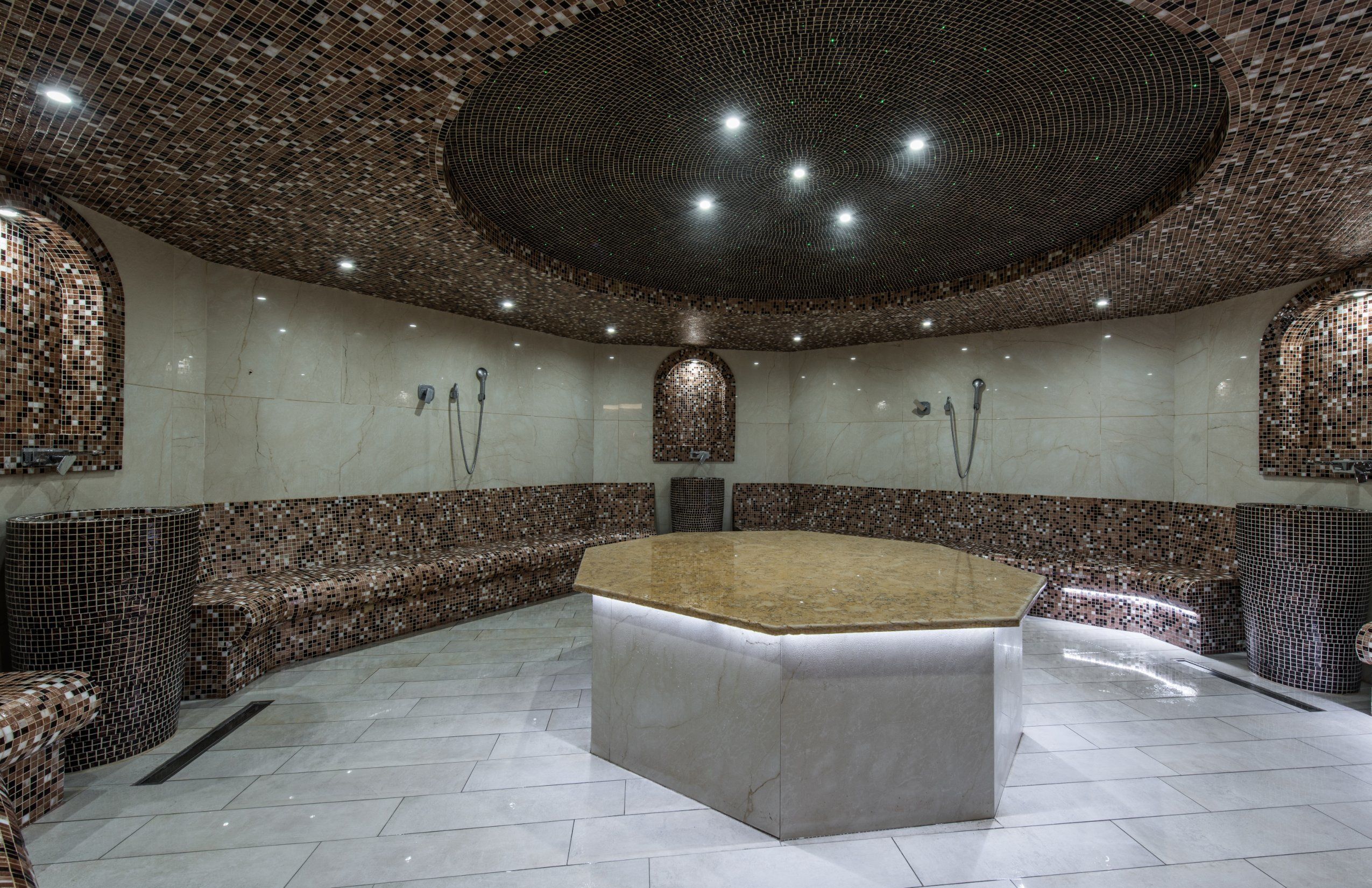 A spacious, well-lit room with a central marble-topped table, surrounded by seating areas and a decorative mosaic ceiling.