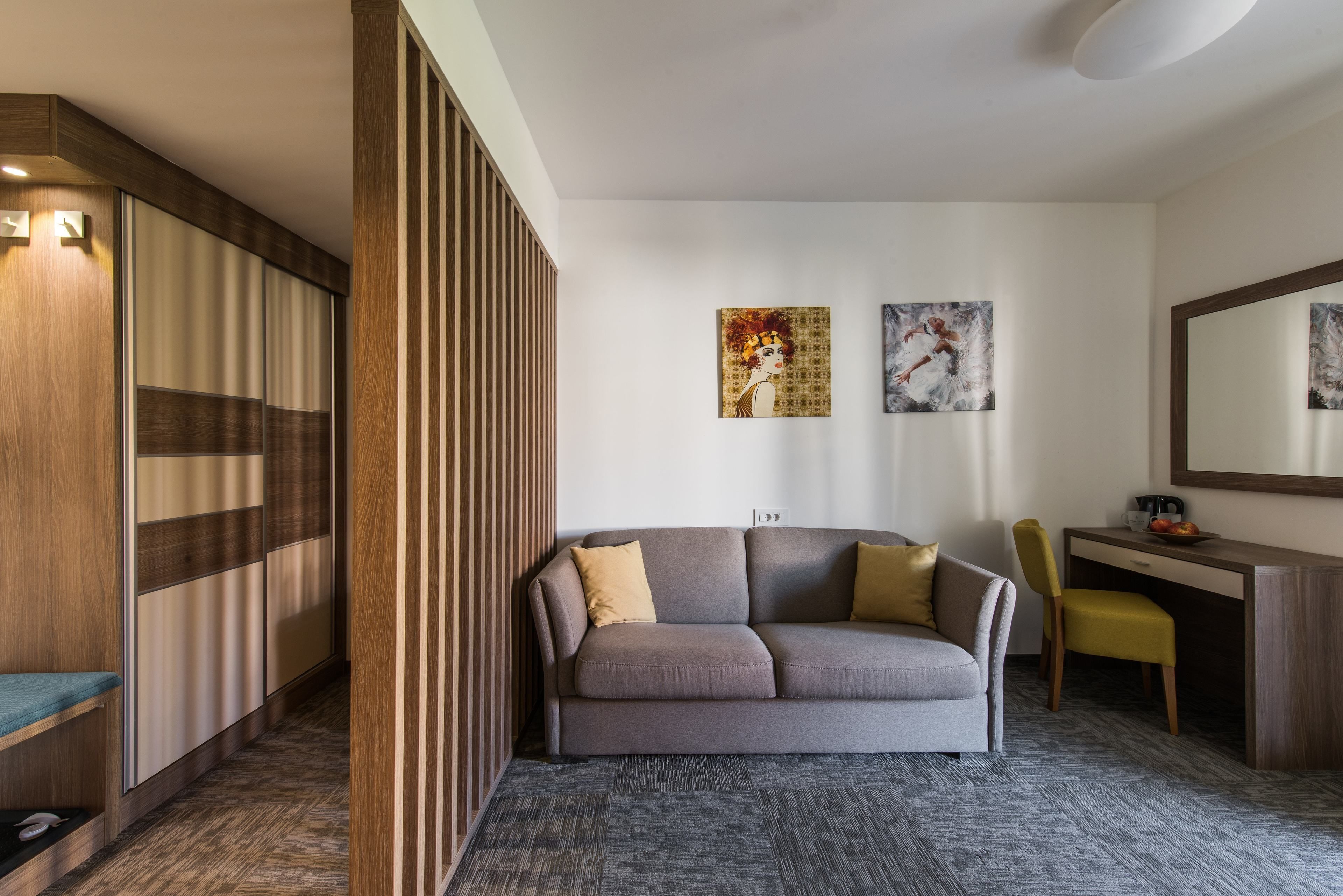 Une chambre d’hôtel moderne, équipée d’un canapé, d’un bureau et d’une chaise. Deux tableaux ornent le mur, et un grand miroir est placé au-dessus du bureau.