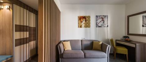 Une chambre d’hôtel moderne, équipée d’un canapé, d’un bureau et d’une chaise. Deux tableaux ornent le mur, et un grand miroir est placé au-dessus du bureau.