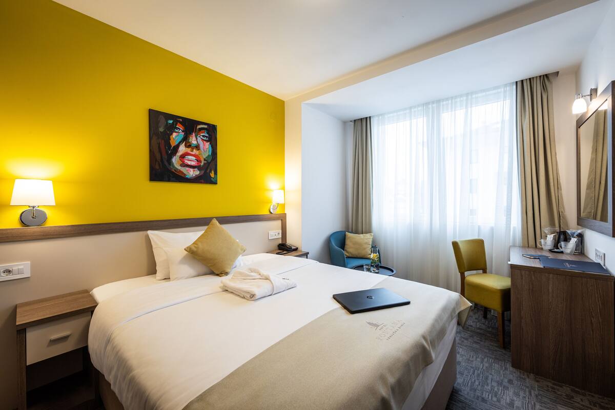 A hotel room with a bed, bedside lamps, a desk, a chair, and a painting on the wall.