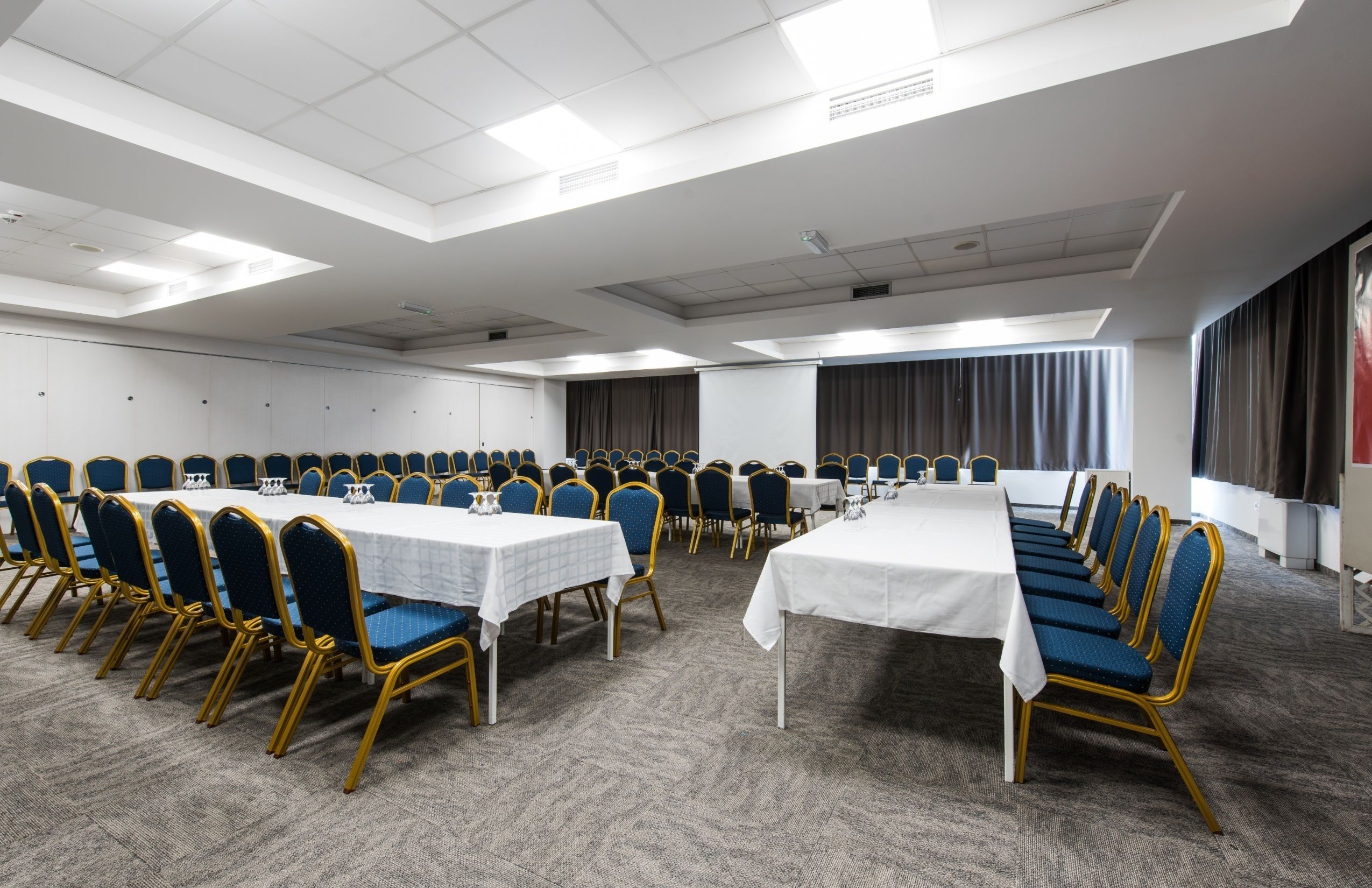 A conference room with multiple tables and chairs, white walls, and a carpeted floor.