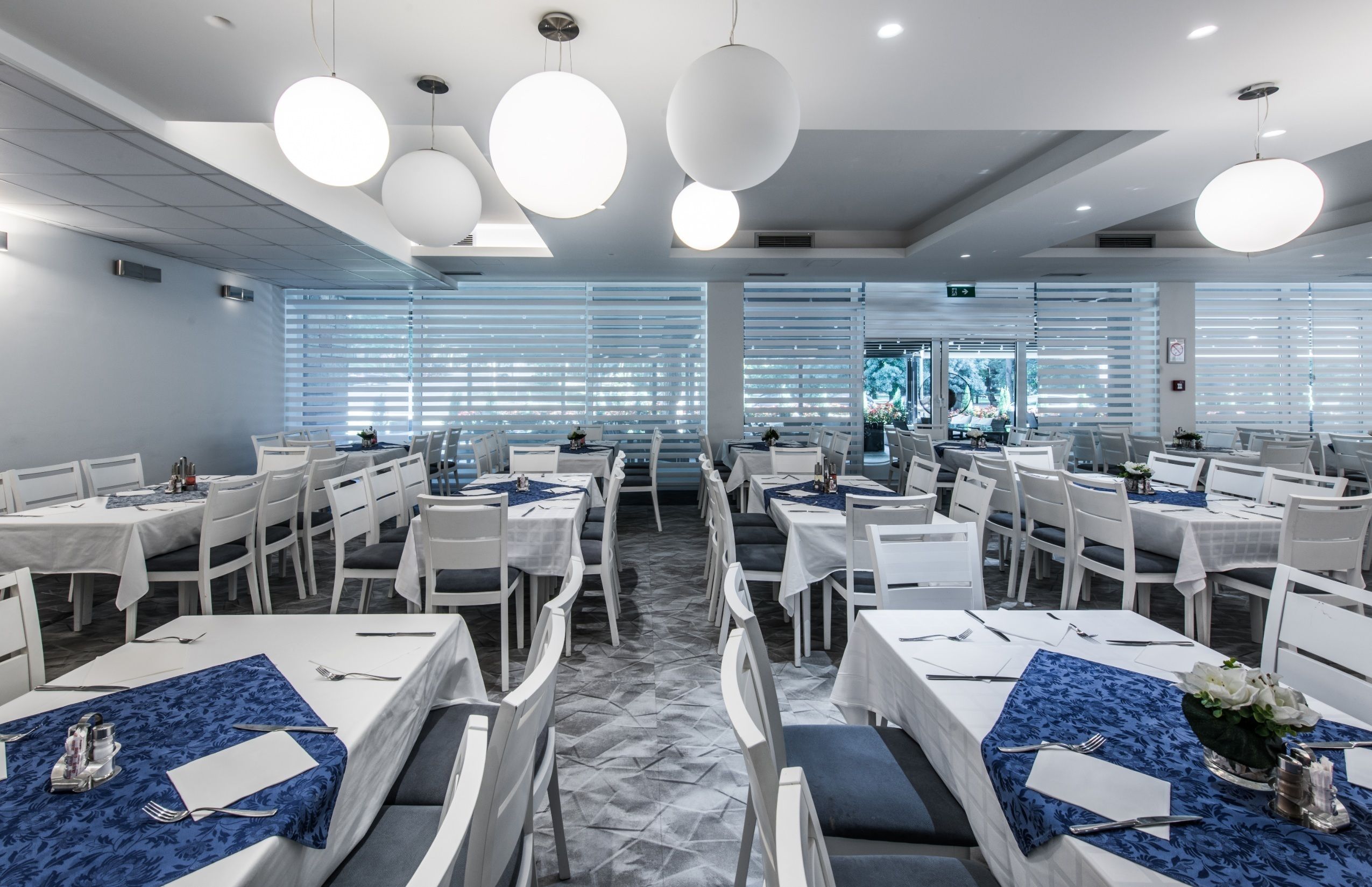 Un comedor moderno con mesas y sillas blancas, manteles azules e iluminación colgante.