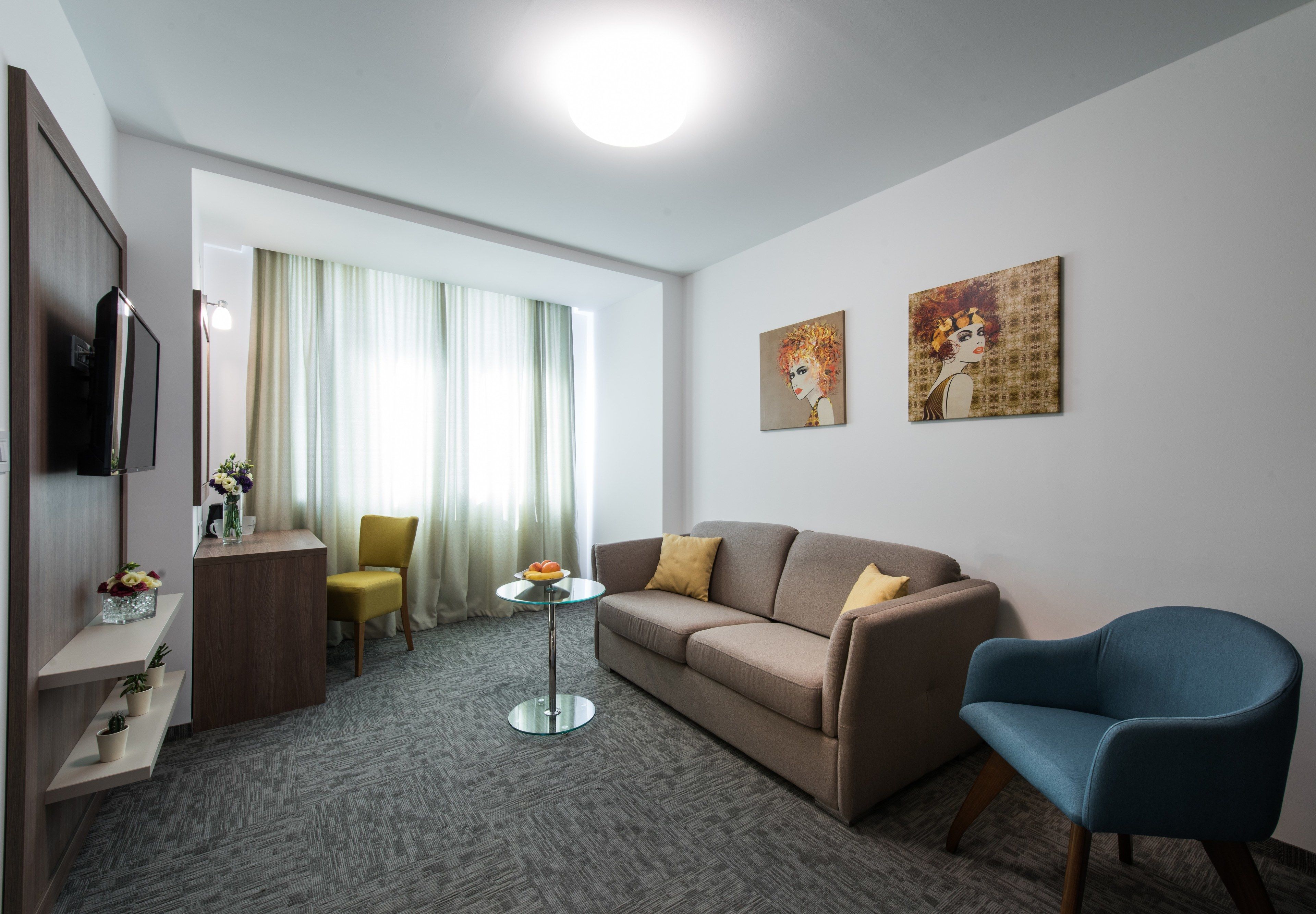 Una habitación de hotel moderna con un sofá, un sillón azul, una pequeña mesa redonda y un televisor sobre un mueble de madera.