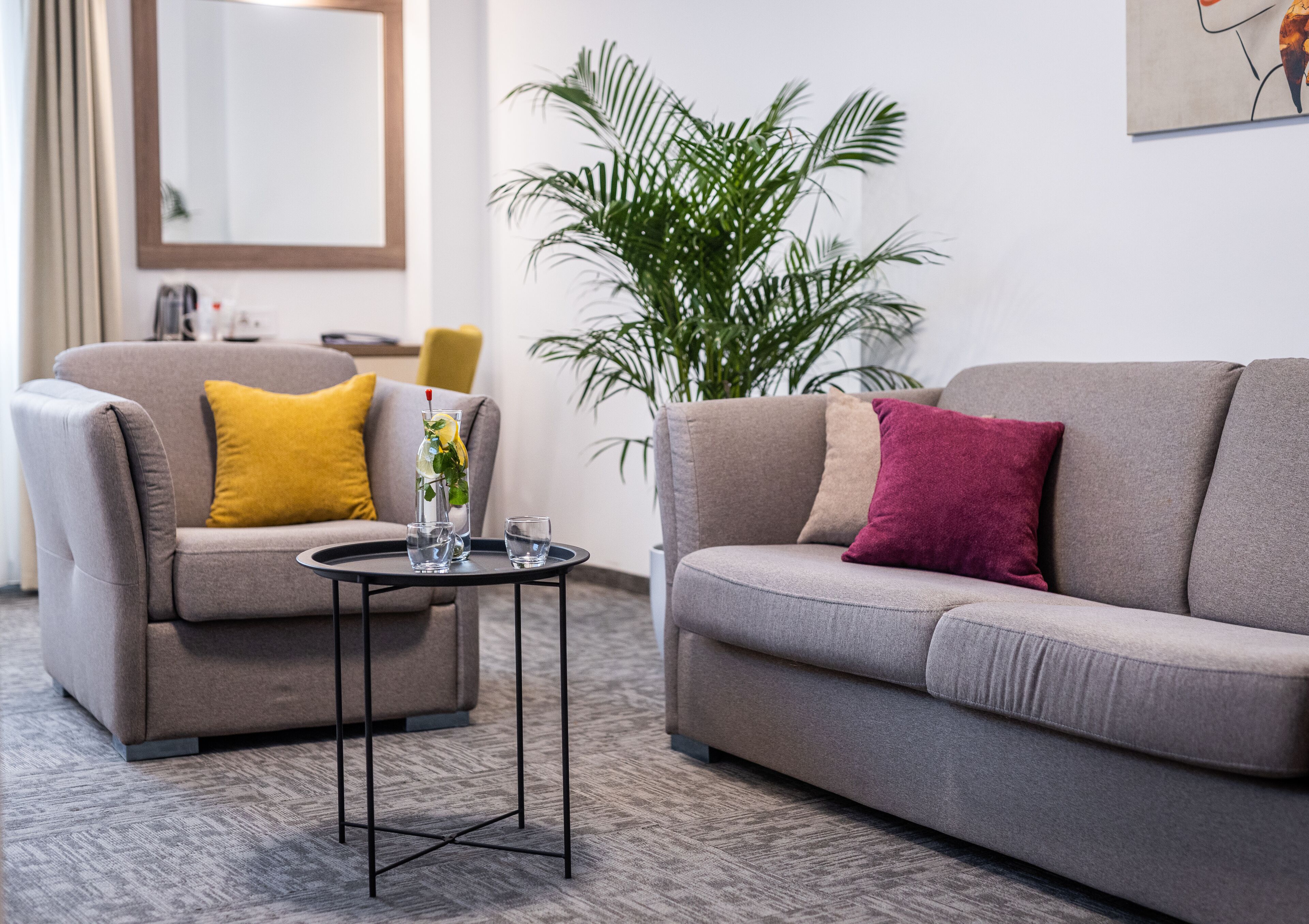 Un salon moderne avec deux canapés gris, l’un orné d’un coussin jaune et l’autre d’un coussin rouge, une petite table basse noire avec un verre et un vase rempli de fleurs, et une grande plante verte en arrière-plan.