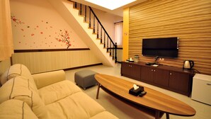 A modern living room with a sofa, ottoman, wooden coffee table, and a flat-screen TV mounted on a wooden panel wall. A staircase with a railing is visible in the background. - Seasons B&B (Liuqiu)