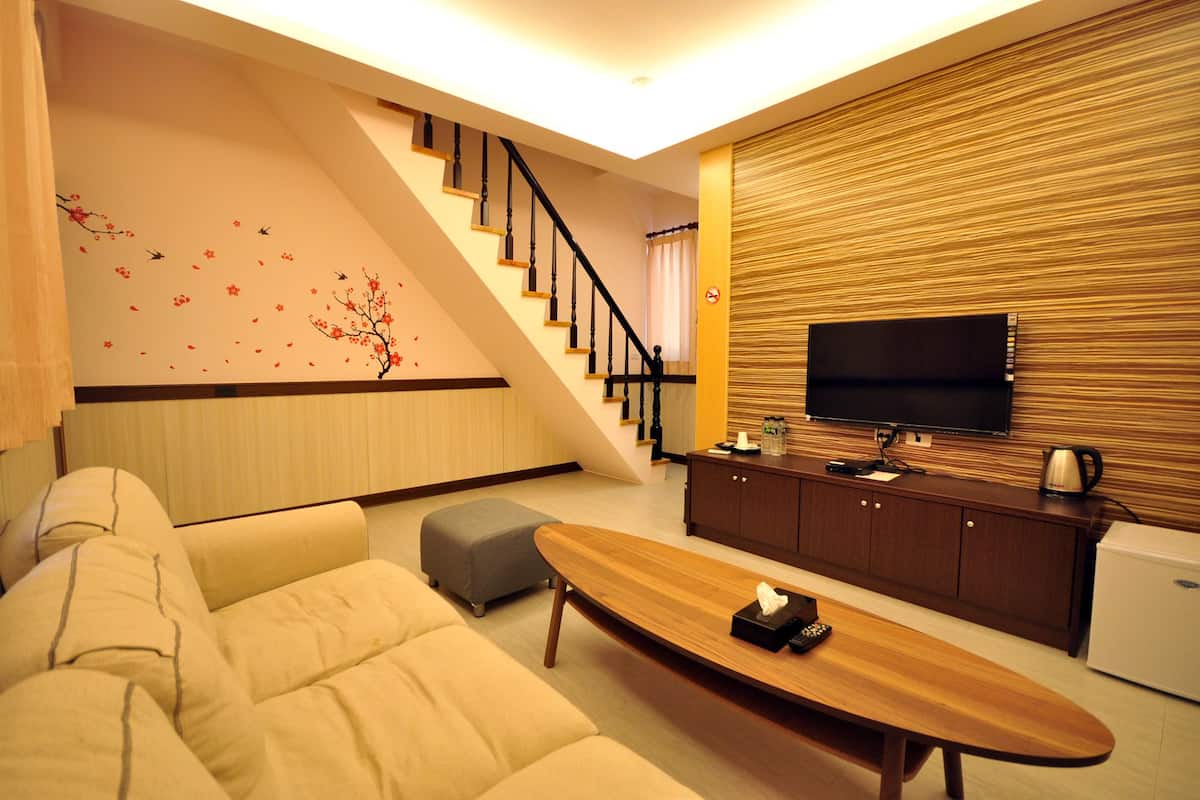 A modern living room with a sofa, ottoman, wooden coffee table, and a flat-screen TV mounted on a wooden panel wall. A staircase with a railing is visible in the background.