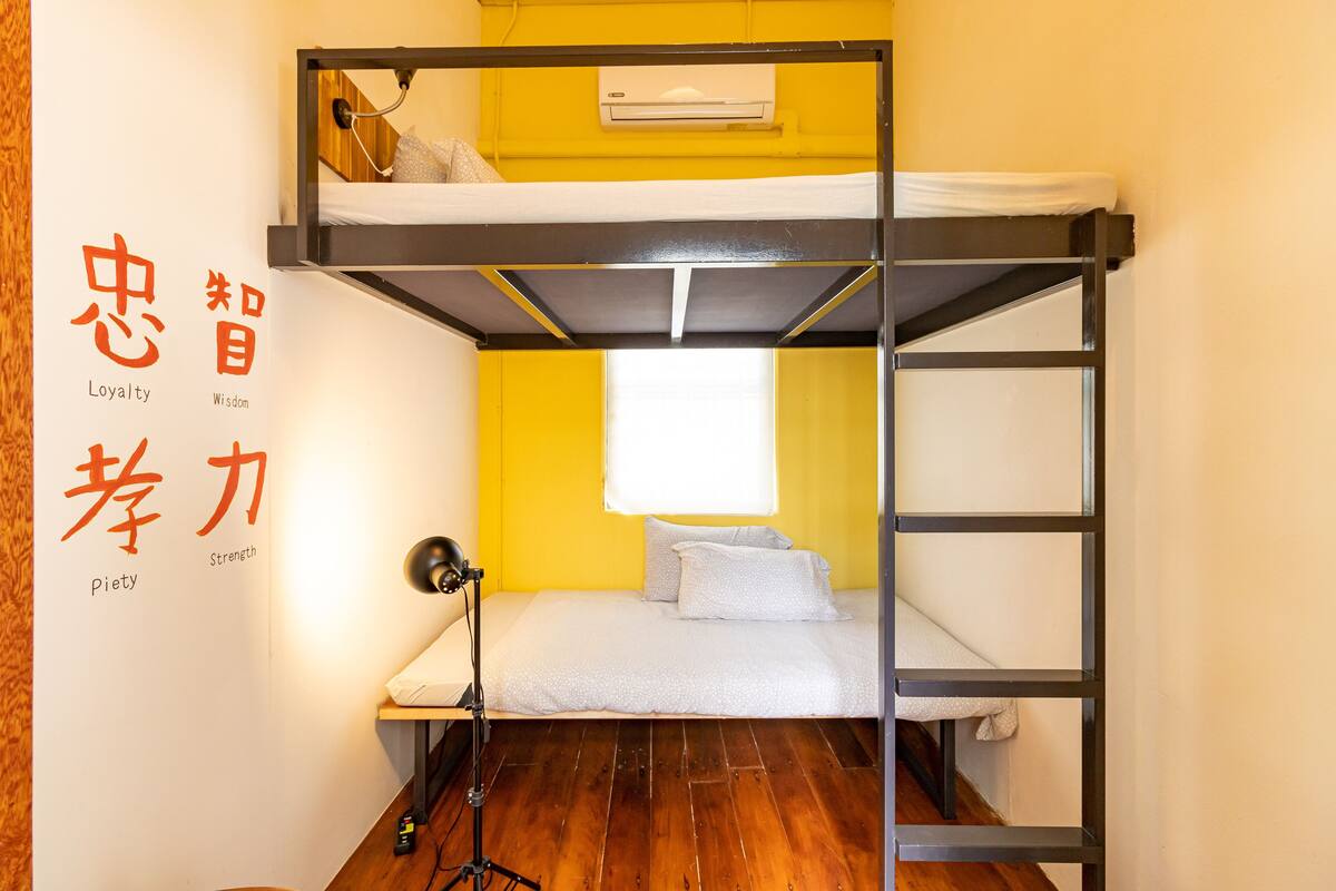 A bunk bed room with a wooden floor, a desk with a lamp, and a window with white curtains.