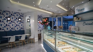 A bakery interior with a display case of pastries, seating area with tables and chairs, and a wall with floral wallpaper.