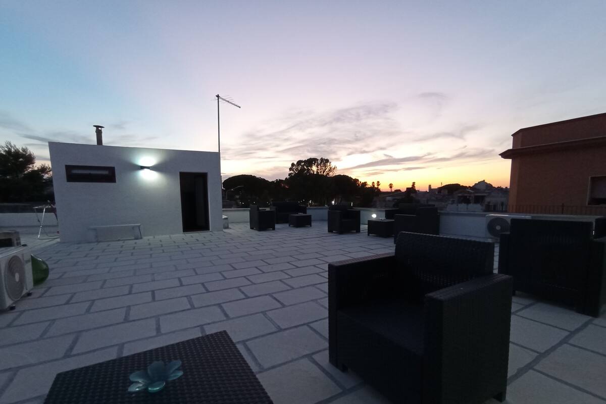 Un espace extérieur aménagé avec des chaises et des tables, un petit bâtiment surmonté d’un drapeau, et un coucher de soleil en arrière-plan.