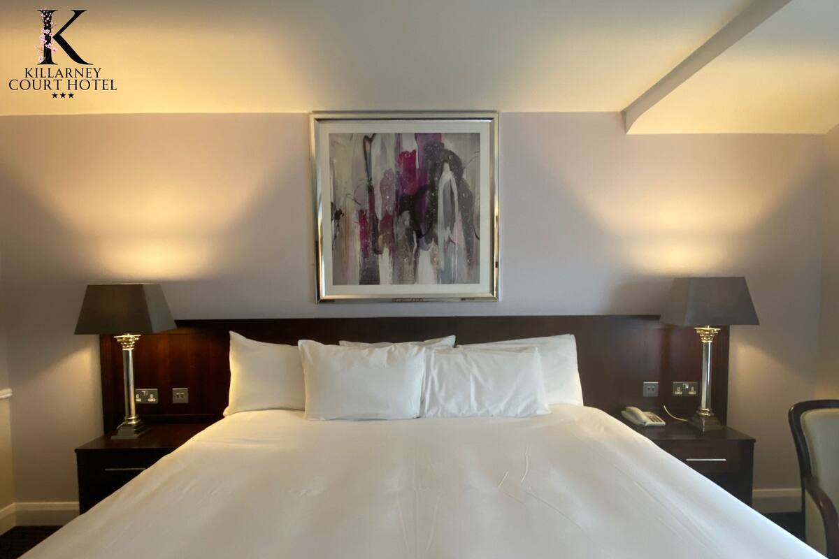 A hotel room with a large bed, two bedside tables with lamps, a framed abstract painting on the wall, and a logo in the top left corner.