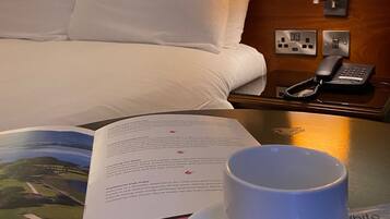 Una habitación de hotel con una cama, una mesita de noche con un libro, una taza y una cuchara.