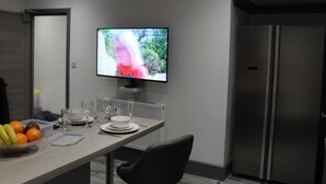 A modern kitchen with a dining area, featuring a flat-screen TV mounted on the wall, a refrigerator, and a table set for two with a bowl of fruit and glasses. - Townhouse @ 282 Walthall Street Crewe (Crewe)