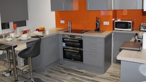 A modern kitchen with grey cabinets, a stainless steel range hood, a microwave, and a toaster. There are two bar stools and a fruit bowl on the counter. - Townhouse @ 282 Walthall Street Crewe (Crewe)