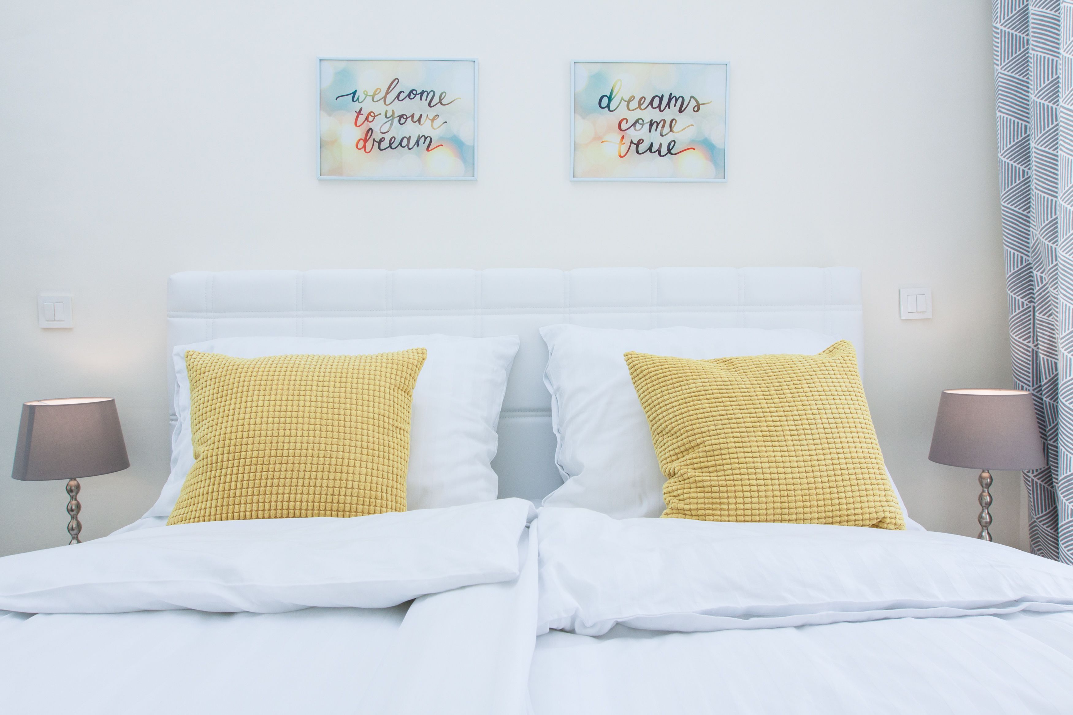 Un letto rifatto con cuscini bianchi e gialli a quadretti, due opere d'arte motivazionale appese al muro e una tenda decorata.