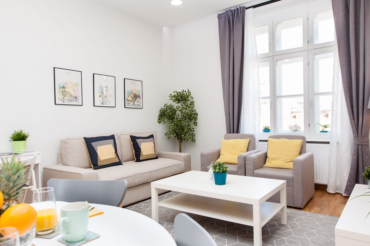 A modern living room with a sofa, armchairs, a coffee table, and a dining area with a table and chairs. There are framed pictures on the wall, a plant, and a window with curtains.
