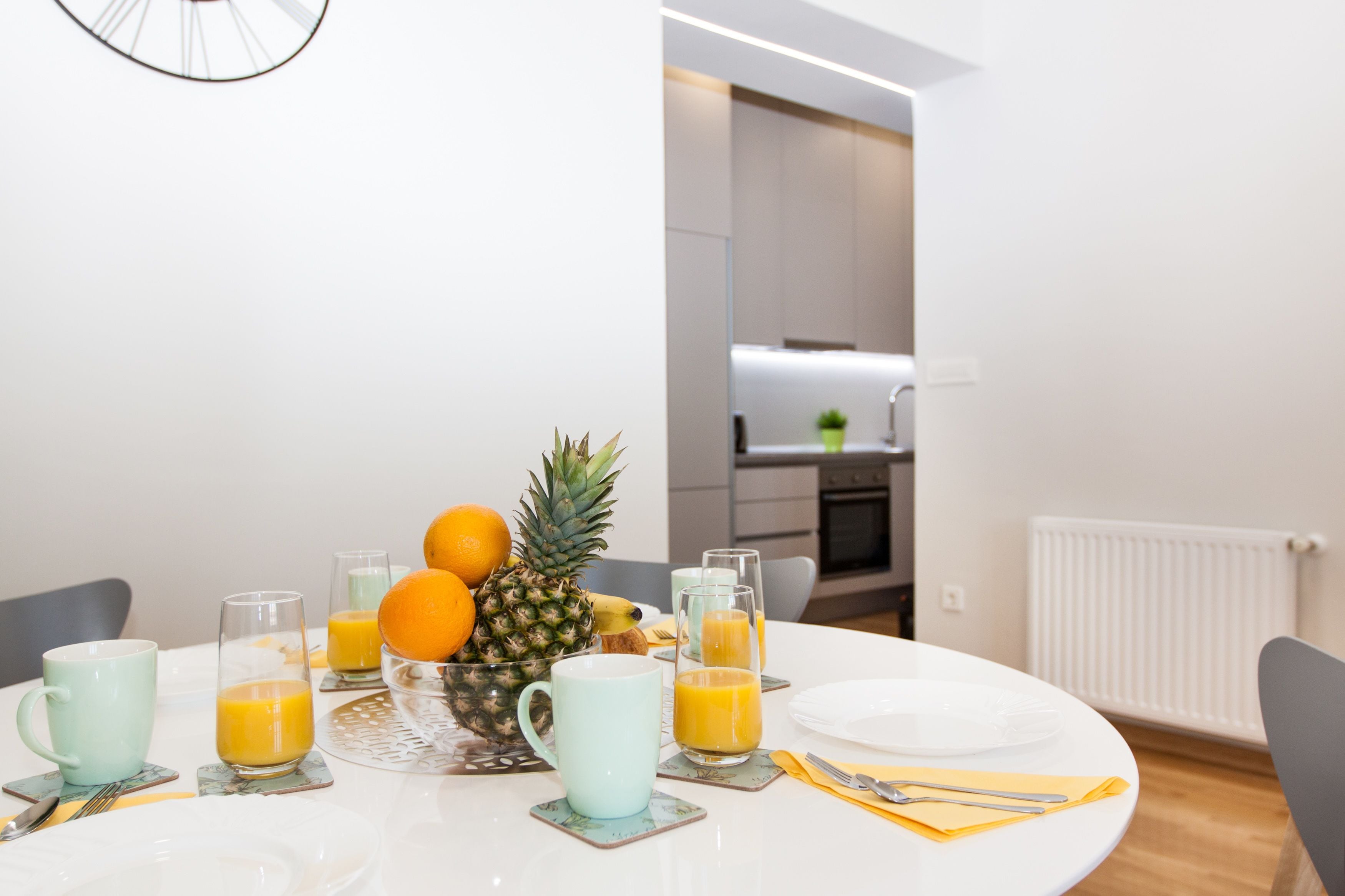 Een ontbijttafel met fruit en sapglazen, een witte tafelkleed en een moderne keuken op de achtergrond.