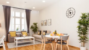 A modern living room with a sofa, coffee table, dining table, and chairs. There is a window with curtains, a wall clock, and framed pictures. - Luxury Main Square Apartments (Zagreb)