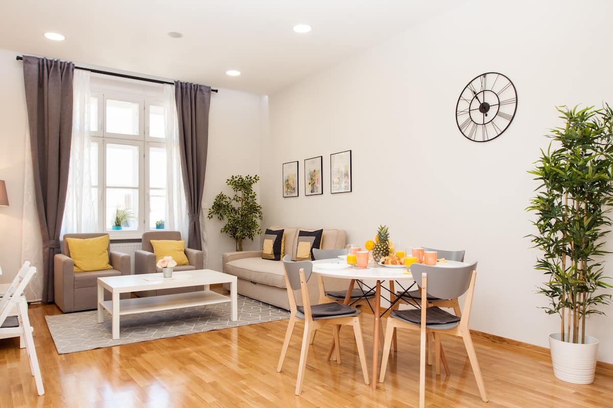 A modern living room with a sofa, coffee table, dining table, and chairs. There is a window with curtains, a wall clock, and framed pictures.