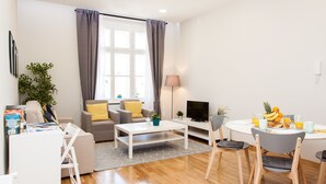 A modern living room with a sofa, coffee table, and dining area with chairs. There is a TV and a plant on a stand. - Luxury Main Square Apartments (Zagreb)