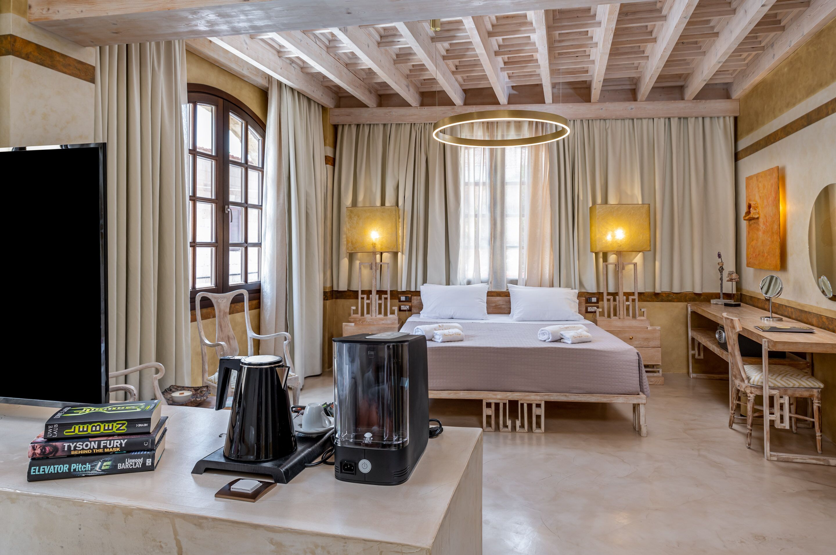 Habitación de hotel con cama, lámparas de noche, cafetera, televisor y escritorio con libros.