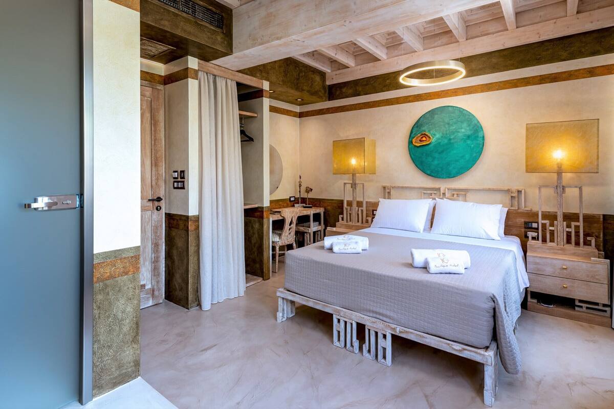 Une chambre d’hôtel avec un lit, des tables de chevet, une grande œuvre d’art circulaire accrochée au mur et un plafond avec des poutres apparentes.