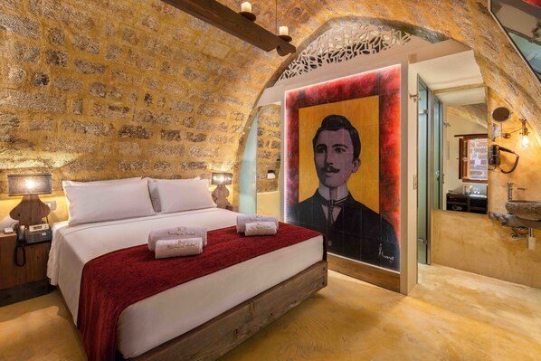 A bedroom with a stone wall, a bed with white linens, a red throw blanket, and a portrait mural.