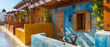A covered outdoor seating area with wooden benches and a glass table, adjacent to a building with a blue facade and wooden doors.