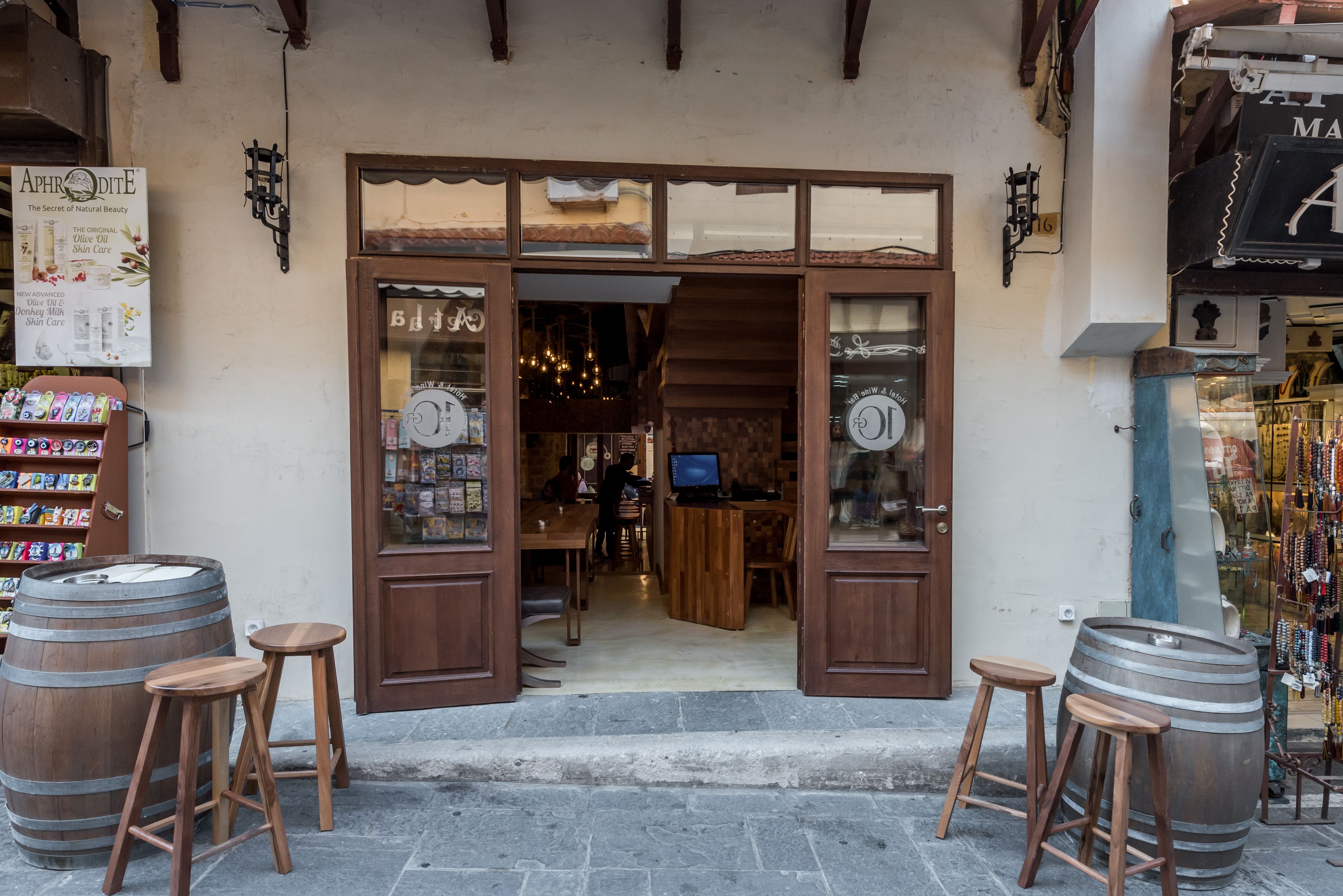 Un local rústico con barriles y taburetes de madera afuera, un letrero anunciando productos de belleza naturales, y una vista parcial del interior con muebles de madera.