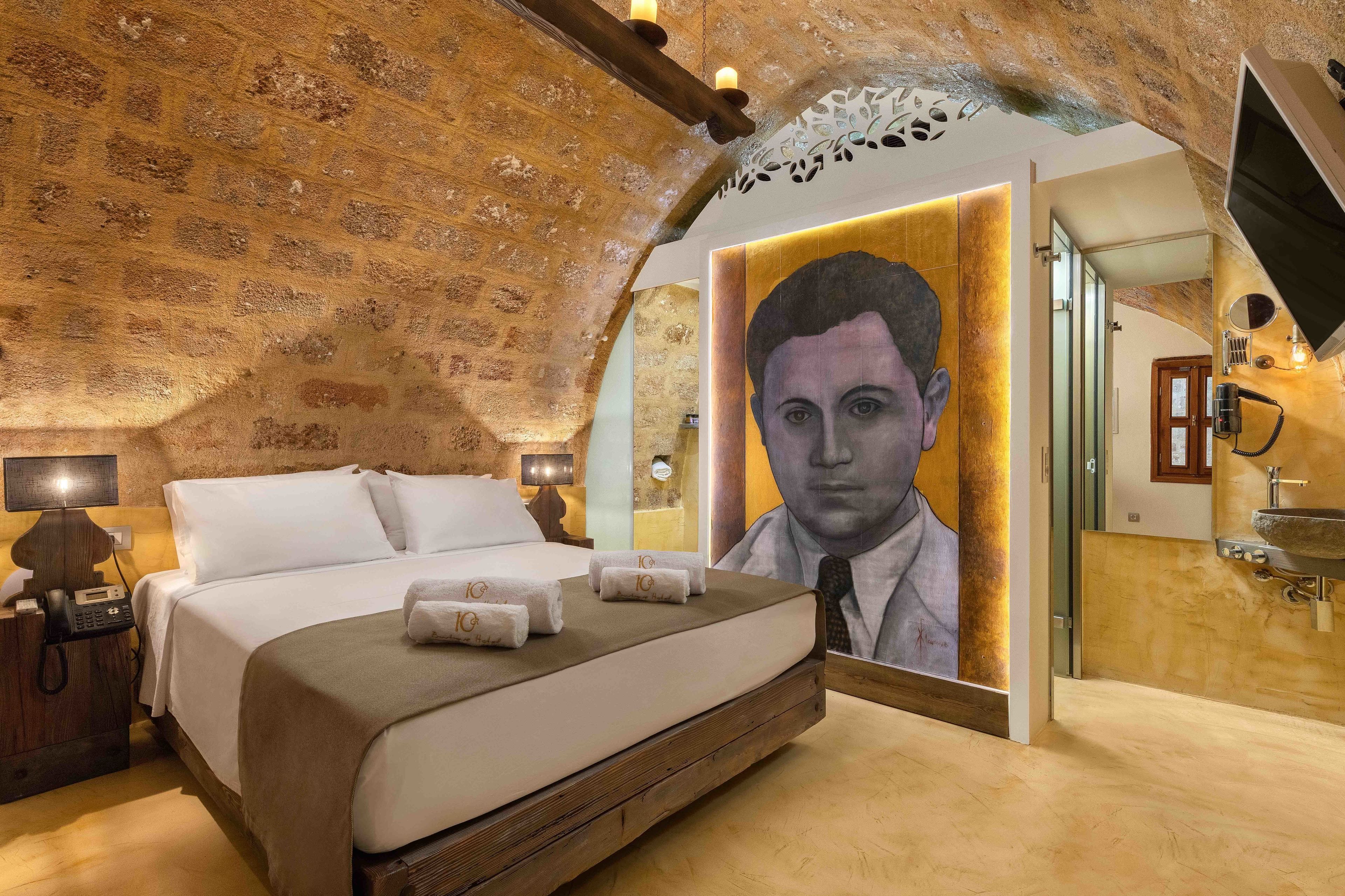 A bedroom with a large bed, a painting on the wall, a bedside table with a lamp, and a bathroom visible through an archway.